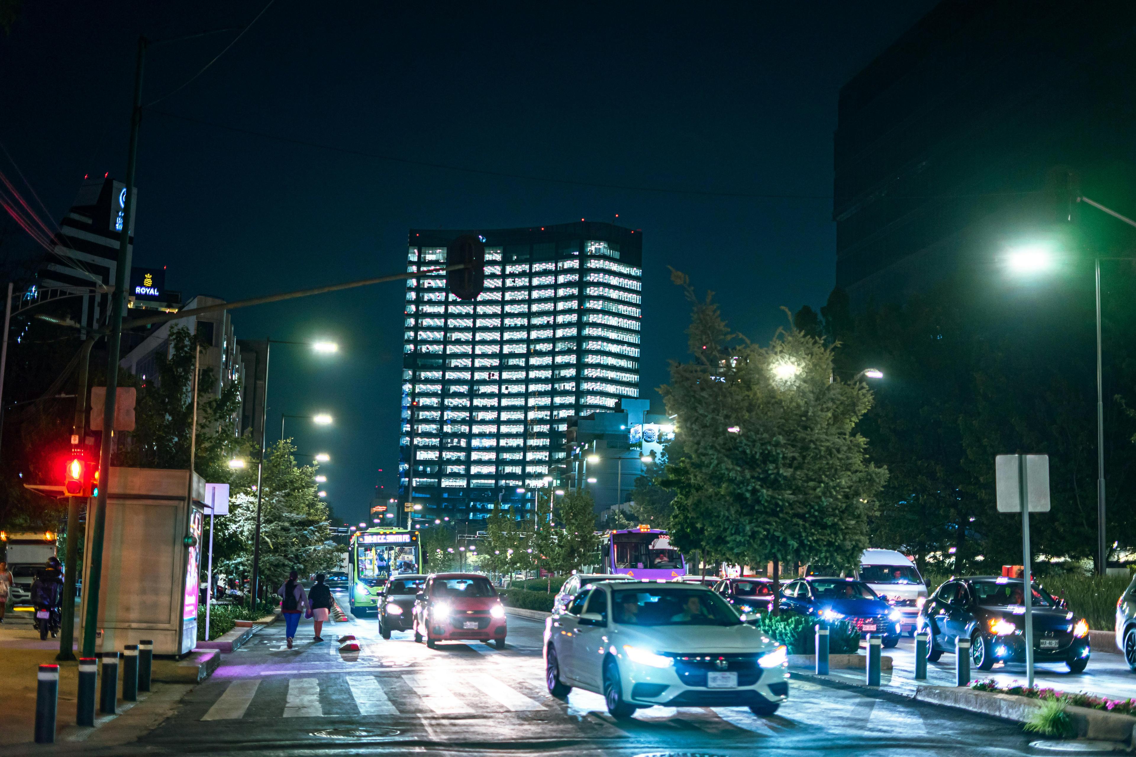 A street scene in Roma Norte, showcasing the iluminated buildings and vibrant streets at night, with pedestrians and cars.